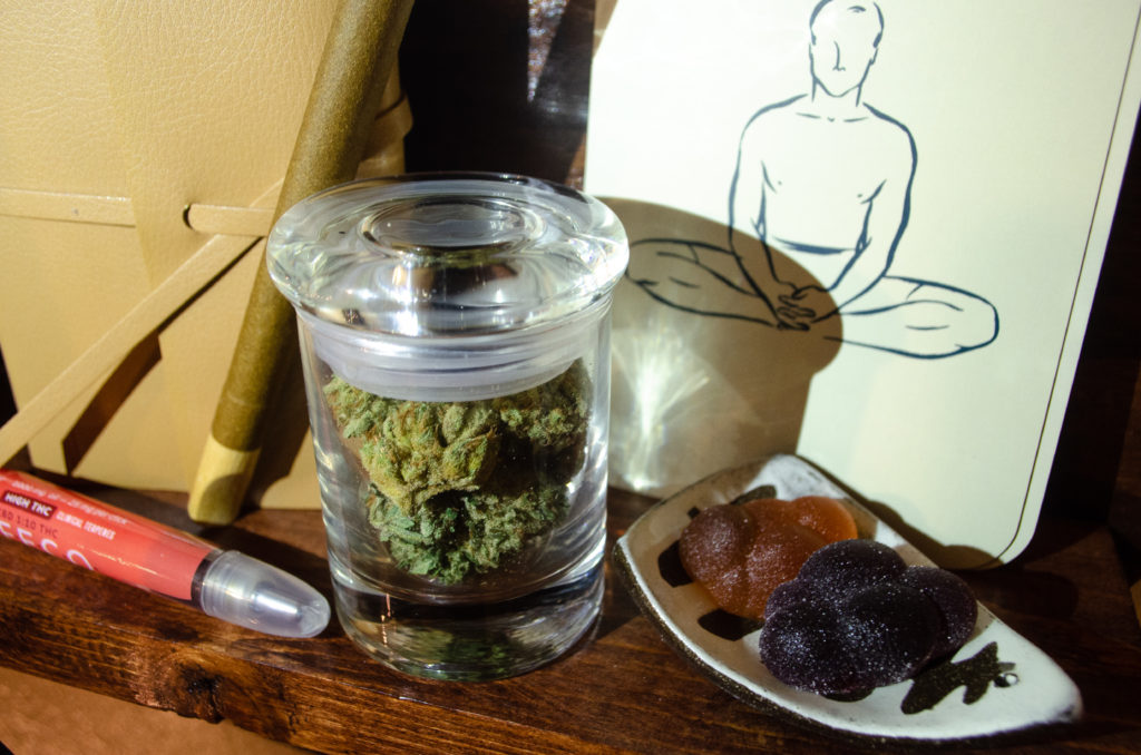Cannabis flower in jar, journal, blunt, cannabis oil, and edibles on dark brown wood shelf to show evening wind-down